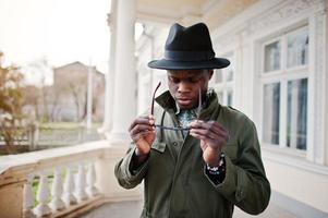 Fashion portrait of black african american man on green coat cloak and black hat, stay on the balcony of yellow mansion photo