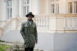 Fashion portrait of black african american man on green coat cloak and black hat, background yellow mansion photo