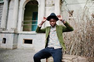 Fashion portrait of black african american man on green velvet jacket and black hat, sitting at felled tree background old vintage house with arch and columns photo