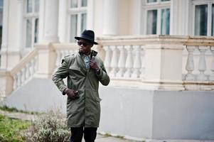 retrato de moda de un hombre afroamericano negro con capa de abrigo verde y sombrero negro, mansión amarilla de fondo foto