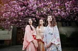 Three european girls wearing traditional japanese kimono background blossom pink sakura tree photo