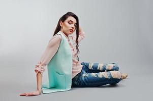 Sitting portrait young brunette girl wearing in pink blouse, turquoise jacket, ripped jeans and cream shoes .Fashion studio shot on gray background photo