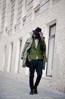 Fashion portrait of black african american man on green velvet jacket, black hat and coat cloak on his shoulders walking on streets of city background house with many windows. Vertical photo