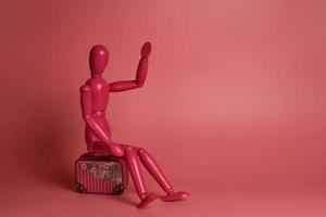 A pink wooden man sits on a suitcase against a pink background. photo