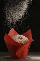 A muffin in red baking paper stands on a table dusted with icing sugar. photo