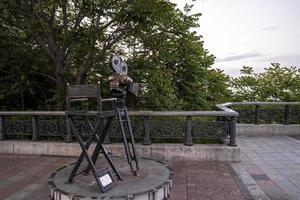 Statue of antique movie camera stands on tripod beside chair with clapboard photo