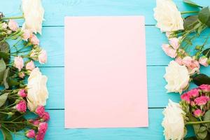 Frame of colorful roses on blue wooden background with paper card and copy space. Top view and selective focus. Mock up photo