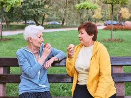 two mature women eating apples on the bench after doing sport exercises in the park. healthy lifestyle concept