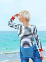 beautiful young blonde caucasian woman on vacation in striped blouse, sneakers and denim overall on the beach by the amazing blue sea background. Happy to be at the sea photo