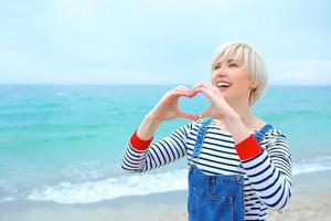 Beautiful young blonde caucasian woman with hands as love sign on vacation. Woman in striped blouse, sneakers and denim overall on the beach by the amazing blue sea background. Happy to be at the sea photo