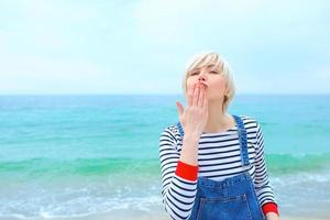 beautiful young blonde caucasian woman on vacation in striped blouse, sneakers and denim overall jumping on the beach by the amazing blue sea background photo