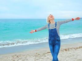 beautiful young blonde caucasian woman on vacation in striped blouse, sneakers and denim overall on the beach by the amazing blue sea background. Happy to be at the sea