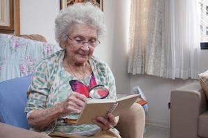 Elderly Lady at home sitting in a chair reading a Book using a Magnifying Glass photo