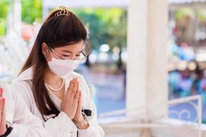 Young woman wearing face mask is raising her hands while listening to sermon from monks. Beautiful etiquette expression. Person wearing white shirts. photo