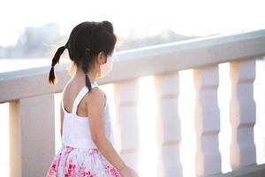 Rear back view of cute young girl walking on bridge. Evening sunlight reflects on water surface. A 5 year old kid is wearing face mask. During summer or spring time. photo