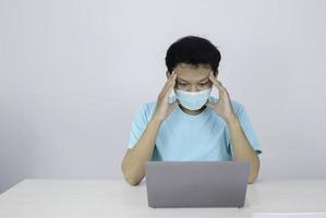 Young Asian man wearing medical mask is feeling unhealthy, tired, and confused with working in laptops on the table. photo