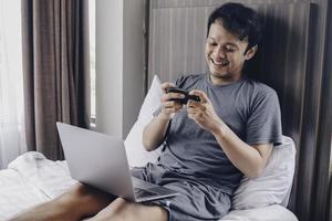 Young Asian man is happy and enjoy when playing a game in laptop and phone at bed photo