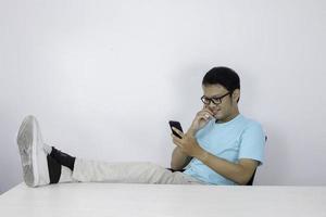 Young Asian Man with happy face with what he see on a mobile phone with leg on the table. Indonesian man wearing blue shirt. photo