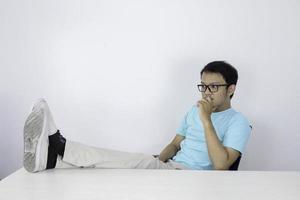 joven asiático con mirada seria y enfocada con la pierna sobre la mesa y el brazo en la barbilla. hombre indonesio con camisa azul. foto