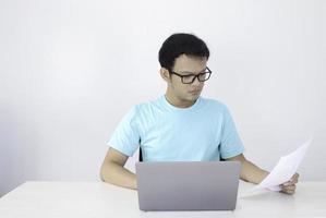 el joven asiático es serio y se enfoca cuando trabaja en una computadora portátil y documenta sobre la mesa. hombre indonesio con camisa azul. foto