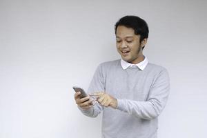 joven asiático conmocionado y feliz con lo que ve en el teléfono inteligente en un fondo gris aislado. foto