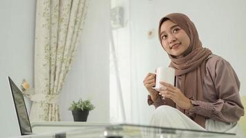 asian woman in hijab working from home using laptop while enjoying a drink photo