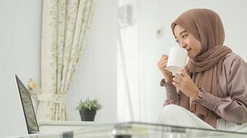 asian woman in hijab working from home using laptop while drinking a drink