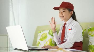 asian elementary school girl studying online at home saying hello to laptop screen photo
