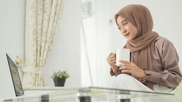 asian woman in hijab working from home using laptop while enjoying a drink photo