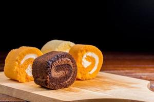 chocolate, vanilla and orange roll cake with whipping cream on  a wooden chopping board and table at the home kitchen with a black background and copy space. Homemade bakery concept. photo