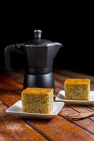 Close up square cut of homemade sweet and solf banana cake on white plate on table with solf focus black moka pot. Delicious and healthy bakery. photo