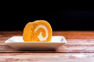 Close up delicious orange roll cake with whipping cream in a square white plate on a wooden table at the home kitchen with a black background and copy space. Homemade bakery concept. photo