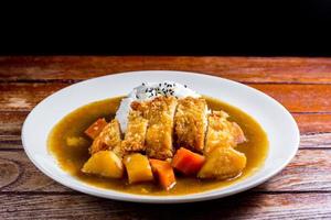 Japanese curry with rice, black sesame, carrot, potato and deep fries chicken in white dish on wooden table. photo