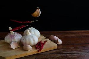 Group of garlic on chopping board and some garlic cloves floating in the air and red dried chilli on wooden table with black background. Copy space for your text.