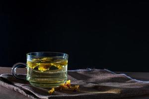 Cup of Chrysanthemum tea with dry flower on brown table cloth on black background. Healthy beverage for drink. Herbs and medical concept. photo