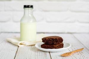 Piece of brownie cake on white small plate with bottle of fresh milk on wooden table and with brick wall with copy space.