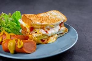 American breakfast concept. Sandwich with melting cheese and blurry sausage and fresh lettuce on a blue plate on a dark rock table. Homemade food in the morning. photo