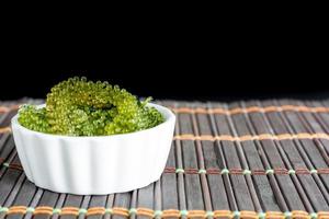 Fresh sea grapes or caviar seaweed in white bowl on wooden mat and black background. Healthy food concept. photo