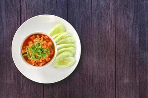 Thai Northern Style Pork and Tomato Relish in white bowl served with cucumber sliced on dark wooden background. Hot and spicy dipping. Top view. photo