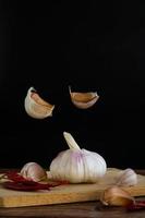 Group of garlic on chopping board and some garlic cloves floating in the air and red dried chilli on wooden table with black background. Copy space for your text. photo