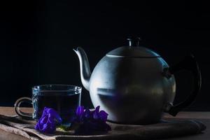 Set of Cup and metal pot of Butterfly pea tea with fresh violet flower on brown tablecloth and wooden table on black background. Healthy beverage for drink. Herbs and medical concept. photo