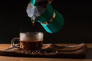 vierta café negro caliente de la cafetera moka verde para limpiar la taza de café con humo y granos de café sobre un mantel marrón y una mesa de madera. beneficio del concepto de café. foto