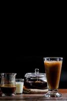Iced milk tea in tall glass with solf focus of transparent teapot with hot tea on brown cloth on wooden table on black background with copy space. Famous beverage of Asian. Healthy drink concept