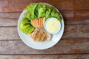 Top view grilled chicken breast steak with sliced avocado, red apple and green lettuce leaves served with salad dressing in white bowl on dish in cafe and restaurant. photo