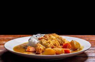 Japanese curry with rice, black sesame, carrot, potato and deep fries chicken in white dish on wooden table. photo