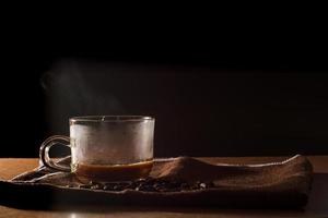 taza de café caliente con humo y un grupo de granos de café sobre un mantel marrón con fondo negro con espacio para copiar el texto. beneficios del concepto de café. foto