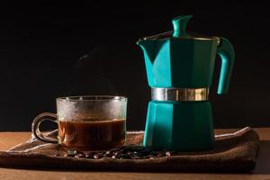 Close up hot of black coffee and green moka pot and coffee beans on brown table cloth and wooden table. Benefit of coffee concept. photo