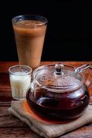 tetera transparente con té caliente sobre tela marrón con un vaso de leche sobre una mesa de madera sobre fondo negro con espacio para copiar. famosa bebida de asia. concepto de bebida saludable foto