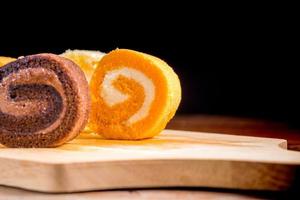 Close up delicious orange roll cake with whipping cream on  a wooden chopping board and table at the home kitchen with a black background and copy space. Homemade bakery concept. photo