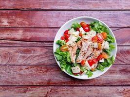 Healthy food concept. Grilled chicken breast salad with fresh red tomato, green lettuce, and mozzarella cheese on a white dish on wooden table. photo